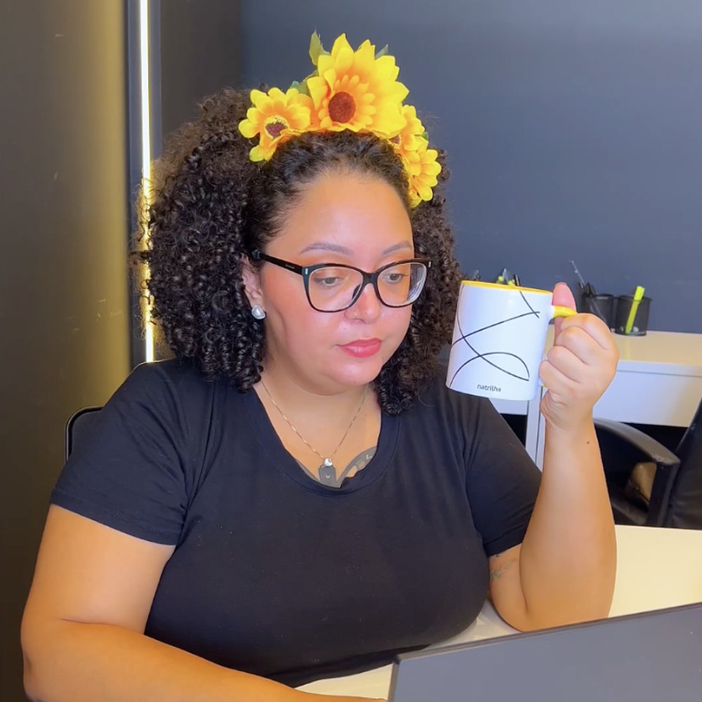 Jennifer com uma coroa de girassóis, segurando uma caneca do Natrilha em frente ao computador.